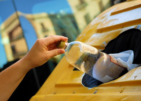 A person wearing a beige sleeve is holding a clear plastic bottle with a green cap, positioned above a yellow waste bin attached to a vehicle or skip. The bottle appears to contain a transparent liquid, and the bin is partially open, showing some mixed waste material and plastic packaging inside. The scene is set outdoors, with a blurred background of buildings and blue sky, indicating a typical rubbish collection or disposal environment. The image highlights the process of waste handling, fitting within the scope of rubbish removal services offered by companies such as Office Clearance Richmond upon Thames in the local Richmond area.