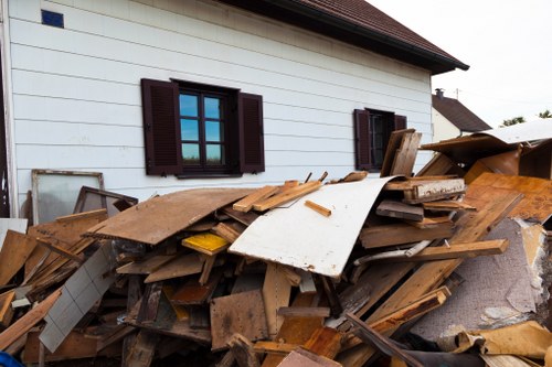 A large pile of discarded wooden and cardboard debris is stacked against the exterior white wall of a house, with dark brown window shutters visible. The rubbish includes various lengths and thicknesses of timber, some with rough, splintered textures and others smoother, alongside pieces of cardboard and a white panel. The debris covers part of a paved driveway or pathway, with the side of the house featuring a sloped roof and a second window partially obscured by the pile. The scene appears to be in a residential area within Richmond upon Thames, reflecting ongoing clearance or rubbish removal activities by Office Clearance Richmond upon Thames, suited for interior or exterior rubbish clearance services in the local area.