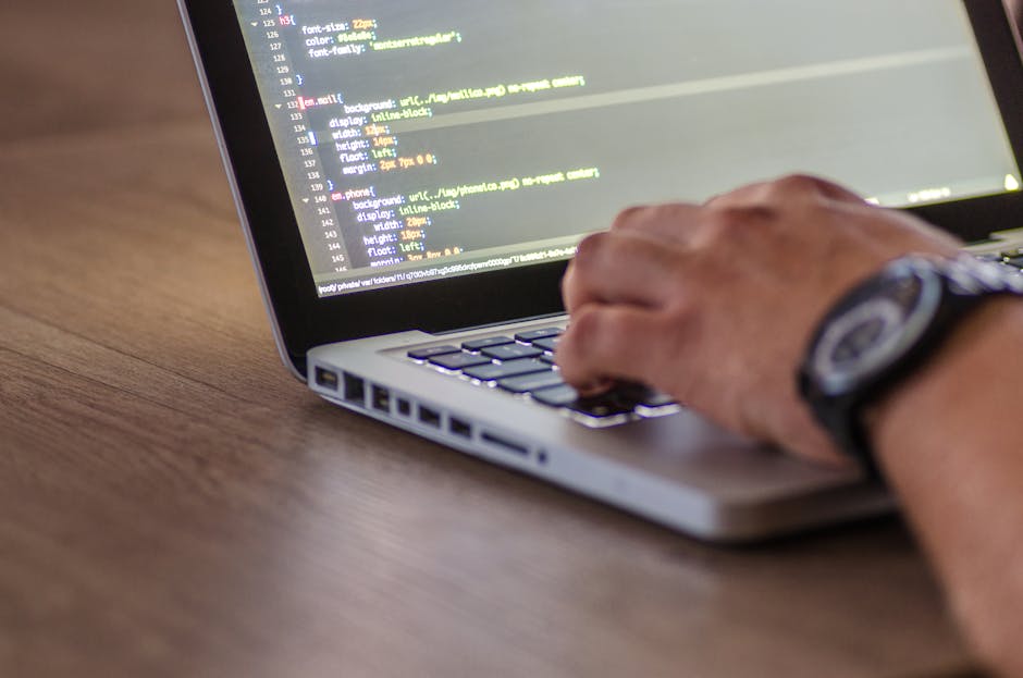 A close-up view of a person's hand typing on a laptop keyboard placed on a wooden desk. The individual is wearing a wristwatch with a dark strap and a round face. The laptop has a silver body with vis