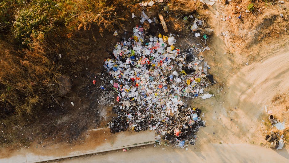 A person wearing a white protective suit, transparent safety goggles, and a black respirator mask stands in front of a large accumulation of mixed waste and rubbish, including plastic bags, cardboard 