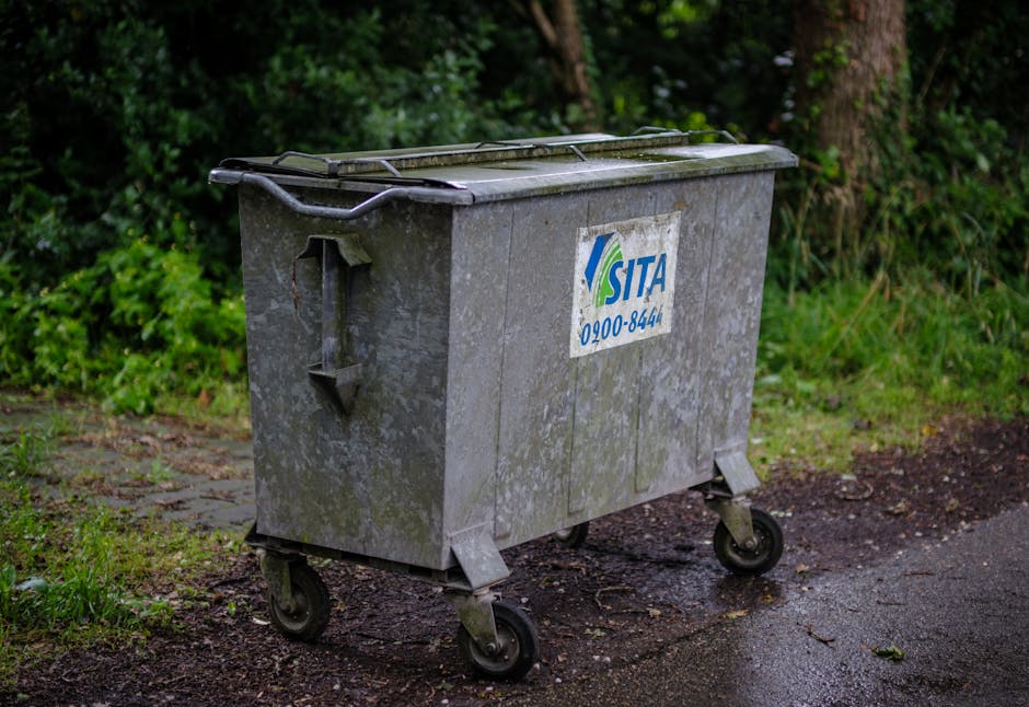 A large, rectangular metal wheeled bin with a hinged lid, positioned outdoors on a paved pathway adjacent to grassy and leafy areas. The bin is made of galvanized steel, showing signs of weathering wi