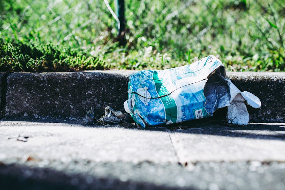A crumpled plastic bag, predominantly white and blue with green accents, lies discarded on a concrete sidewalk, partially resting against the curb. The bag appears to be made of thin, flexible plastic